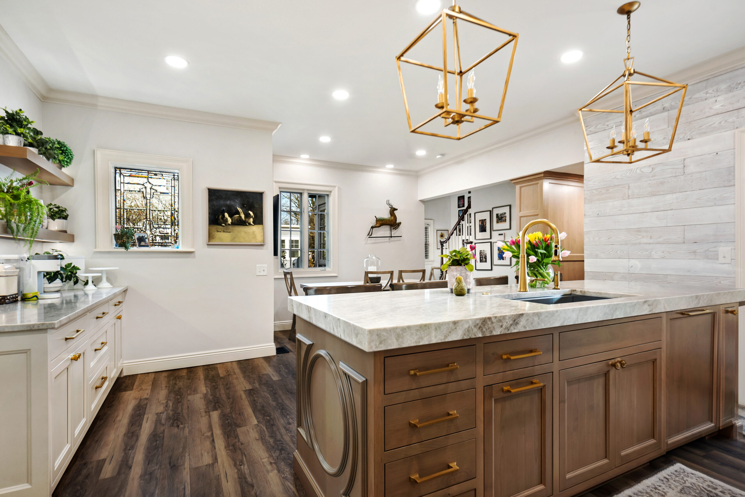 A modern kitchen renovation with wood floors, white and wood cabinetry, marble countertops, gold pendant lights, open shelves with plants, plus a dining area in the background featuring wall art and large windows.