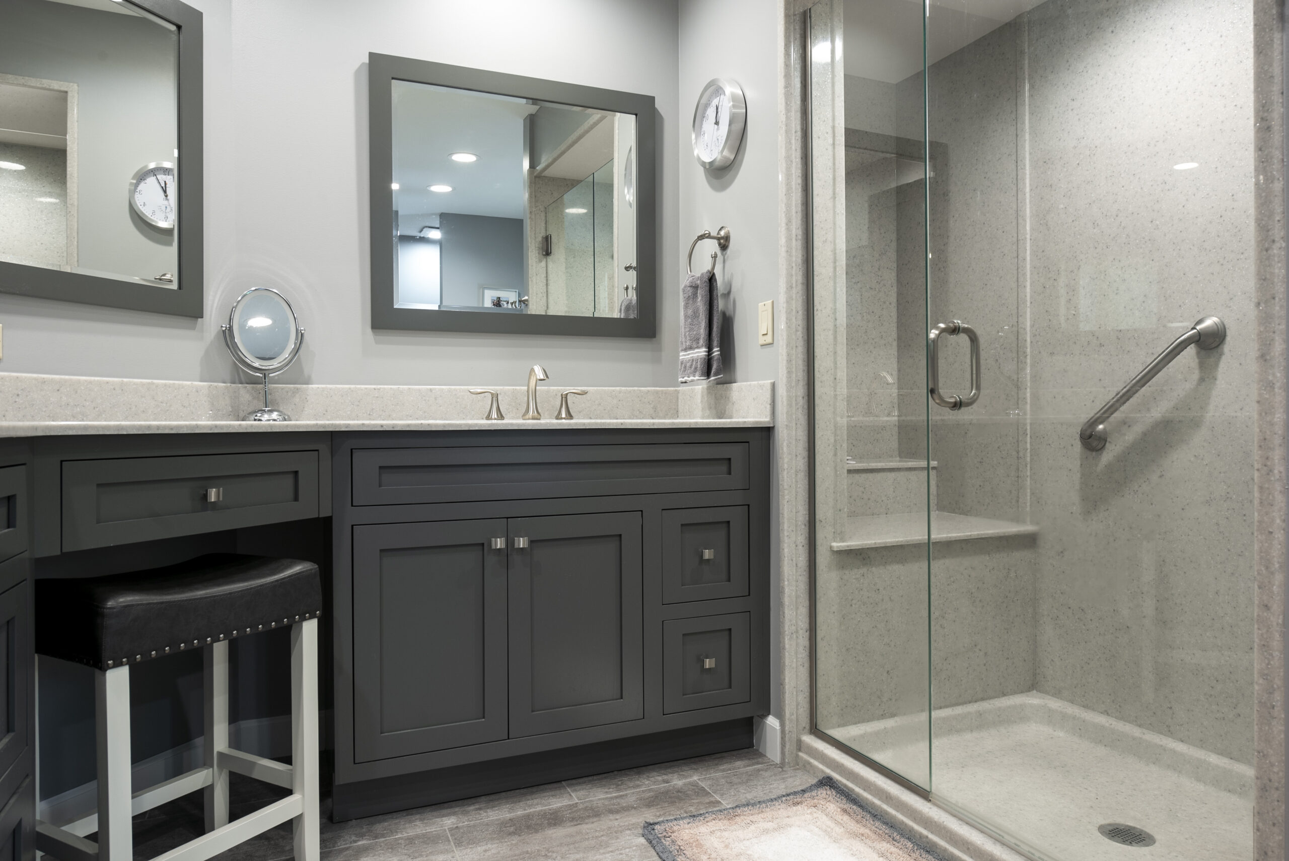 Modern bathroom with dark gray cabinets, a beige countertop, double mirrors, a round wall clock, and a glass-enclosed shower with grab bar and built-in seat. Perfect inspiration for your next bath remodel by a top bathroom remodeling company Columbus Ohio.