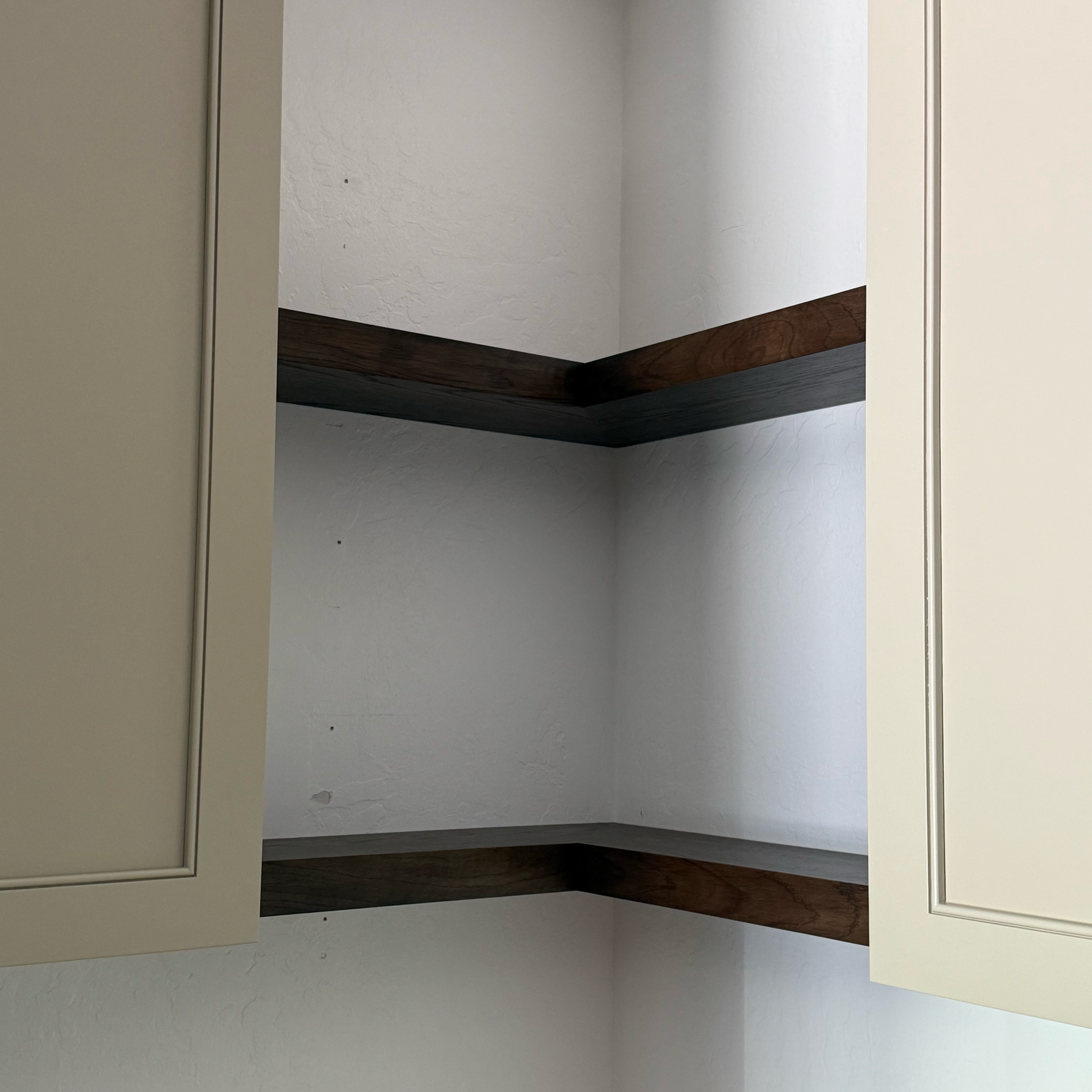 Two beige cabinets with dark wooden corner shelves between them, mounted on a white textured wall.