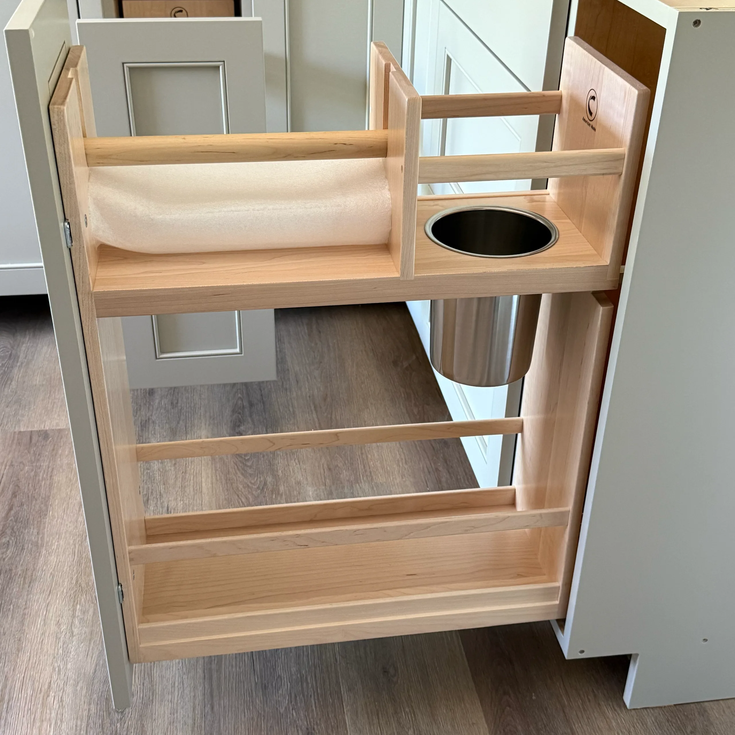 A wooden pull-out kitchen cabinet organizer with two shelf tiers, a paper towel holder on top, and a built-in metal bin, set on a wood floor next to white cabinetry.