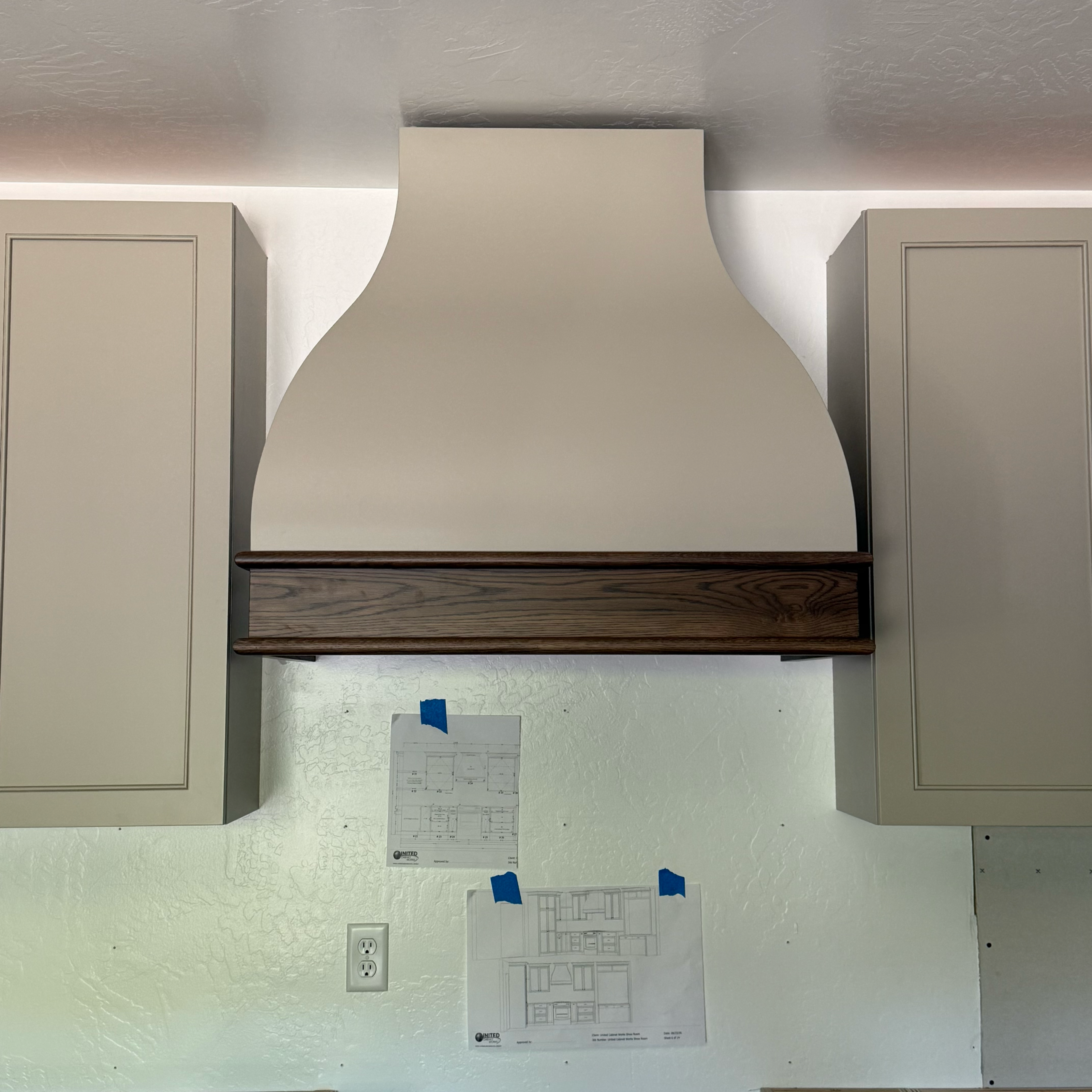 A beige kitchen range hood with a wooden trim is mounted between two cabinets. Below, two papers with architectural drawings are taped to the wall above an electrical outlet.