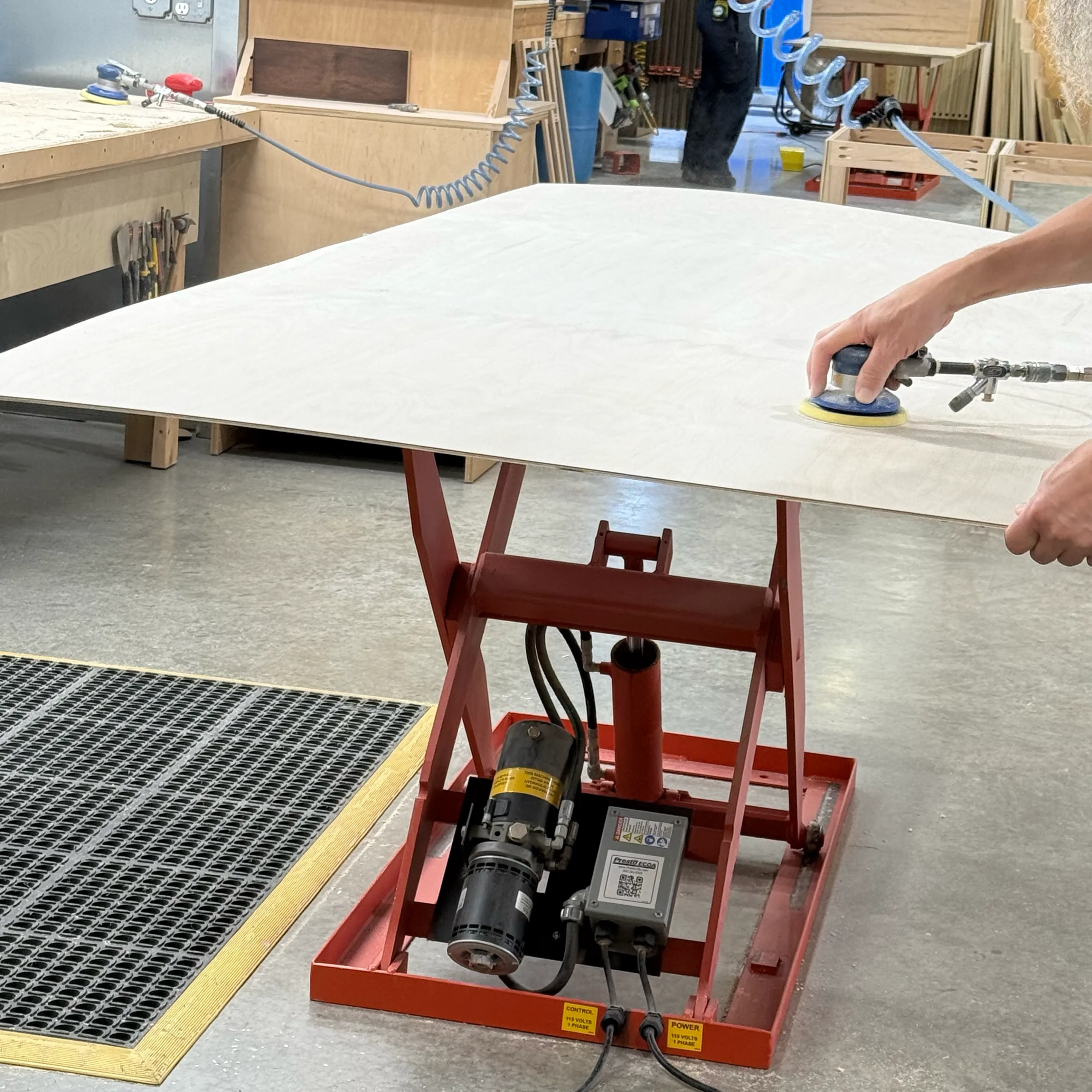 A person uses a handheld power sander on a large sheet of wood placed on a red hydraulic lift table in a woodworking shop.